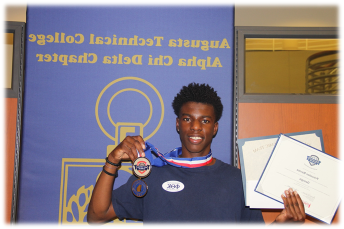 African American male, Princeton Barnes smiles, holding his certificates in his right hand and his medals in his left hand while standing in front of a blue violet poster labelled 奥古斯塔 Technical College Alpha Chi Delta Chapter in gold font. 